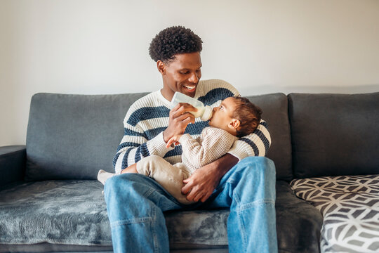 Young father feeding baby with milk bottle on sofa at home