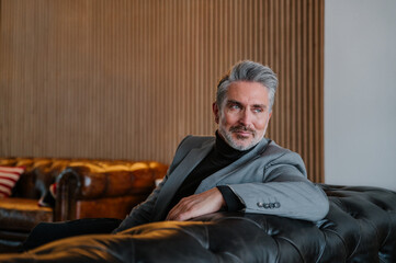 Thoughtful mature man in stylish suit resting on leather couch