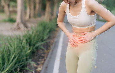 Asian woman experiencing pain in her abdomen during exercise outdoors, wearing white sports bra and light colored leggings, with blurred park background