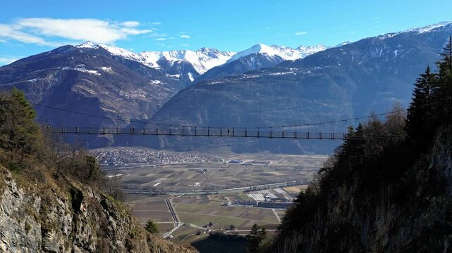 Saillon, Switzerland - 15 February 2025: Tourists Walking Across the Passerelle &agrave; Farinet, a Wooden Bridge Above the Salentse Gorge with Views of the Swiss Alps on a Beautiful Sunny Day