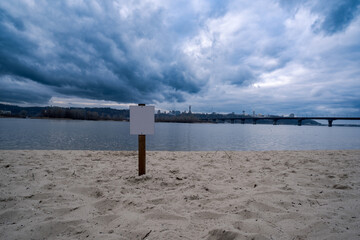 Stormy skies over the Dnipro River in Kyiv with an empty warning sign in the sand: thin ice warning, fishing and swimming ban - Embankment of the Dnipro River in Kyiv