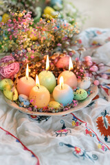 Easter background. Burning candles in shape of colorful eggs and flowers in plate, on table....