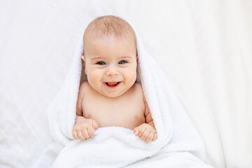 cute little newborn baby with blue eyes on the bed in a white towel after bathing or washing, happy baby at home on a cotton bed smiling