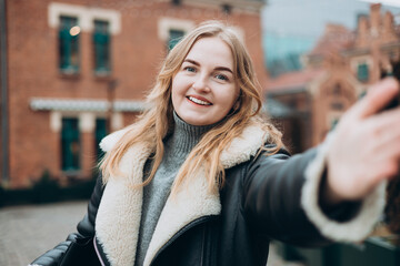 Fototapeta premium Beautiful woman in good mood looks into camera and takes selfie in the street. Blogging, technology, lifestyle, travel, tourism, active life. Winter time