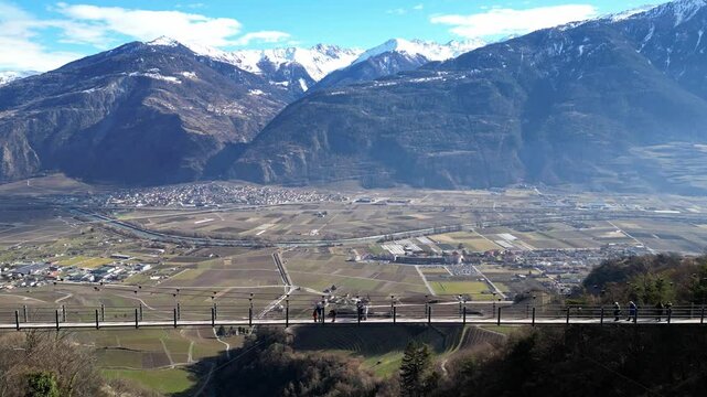 Saillon, Switzerland - 15 February 2025: Tourists Walking Across the Passerelle &agrave; Farinet, a Wooden Bridge Above the Salentse Gorge with Views of the Swiss Alps on a Beautiful Sunny Day