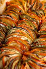 A close-up of a baked ratatouille dish with sliced zucchini, tomatoes, and eggplant arranged in a red ceramic baking dish on a kitchen countertop.