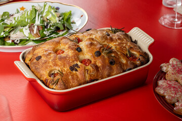 A close-up of crispy focaccia bread with cherry tomatoes, olives, and rosemary in a red baking dish, placed on a red table with a salad and cookies nearby.