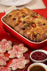 A close-up of crispy focaccia bread with cherry tomatoes, olives, and rosemary in a red baking dish, placed on a red table with a salad and cookies nearby.