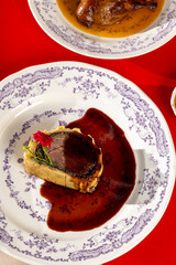 A gourmet beef Wellington slice with a flaky crust, drizzled with red wine sauce, garnished with a red flower, and served on a decorative plate.