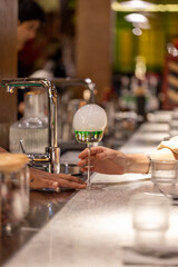 A bartender handing over a molecular cocktail with a green liquid and a smoke bubble on top, served...