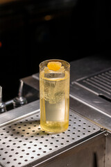 A bubbly golden highball cocktail with a candied ginger garnish, served in a tall glass over ice on a sleek bar counter.