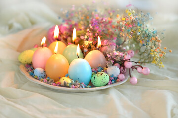 Easter background. Burning candles in shape of colorful eggs and flowers in plate, on table. festive spring seasonal composition. table decor for Easter holiday, Nowruz, Ostara sabbat. soft focus