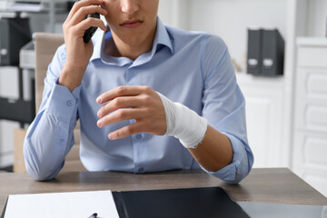 Man with wrist wrapped in medical bandage talking on smartphone at table indoors, closeup