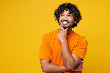Young smiling happy Indian man he wear orange t-shirt casual clothes put hand prop up on chin, lost...
