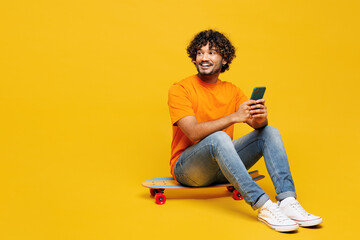 Full body young Indian man he wear orange t-shirt casual clothes sit on skateboard hold use mobile...