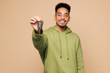 Young man of African American ethnicity in green hoodie casual clothes hold car key fob keyless system stretch hand to camera isolated on plain pastel light beige background studio Lifestyle concept