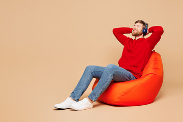 Full body young happy fun man he wear red hoodie casual clothes sit in bag chair listen to calm...