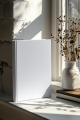 A blank white book mockup standing on a windowsill with soft sunlight casting shadows. A vase with dried flowers is nearby, creating a serene and cozy atmosphere.