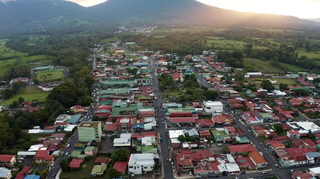 Aerial drone shot trucking left and tilting up revealing the Arenal Volcano overlooking La Fortuna, Costa Rica. Sunset paint the sky, with clouds rolling over the lush landscape and small town below.