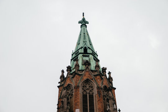 A stunning Gothic church spire with intricate architectural details, aged brickwork, and a patina-covered roof, set against a moody overcast sky