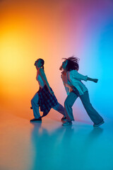 Naklejka premium Dynamic hip hop performance by two young women in casual clothes against gradient studio background in neon light. Concept of movement, breakdance, music culture, artistic expression, youthful energy.