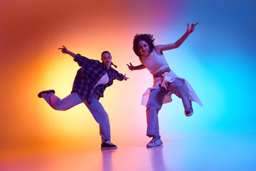 Two young women immersed in energetic hip hop dance against gradient studio background in neon light. Concept of rhythm, street choreography, dynamic expression, breakdance, freestyle movement