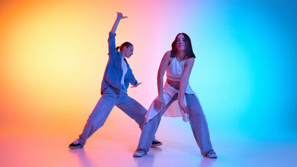 Two young women in stylish casualwear dancing hip hop against neon-lit studio gradient. Concept of rhythm, breakdance, urban culture, freestyle energy, passion for movement, modern dance