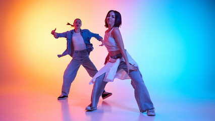Dynamic hip hop energy. Two young women in casual attire dancing against gradient studio background in neon light. Concept of modern dance style, hobby, active lifestyle, youth culture