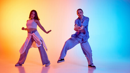 Synchronized hip hop performance by two stylish female dancers against gradient studio background in neon light. Concept of modern dance style, hobby, active lifestyle, youth culture