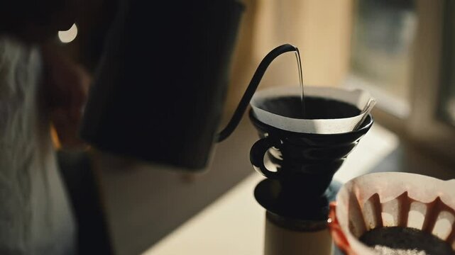 Close-up shot of manual coffee brewing with hot water being poured from a gooseneck kettle into a ceramic pour-over dripper. Morning sunlight shines through the window, creating a calm and cozy atmosp