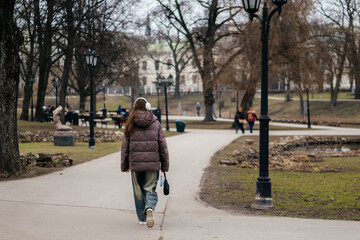 A young woman in a warm jacket and headphones walks through a peaceful city park on a chilly day, surrounded by bare trees and vintage lampposts.