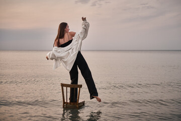 Fototapeta premium Young woman balancing on chair in serene seascape at dusk