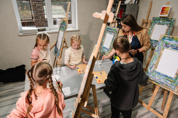 A group of school children with brushes and an easel in art lesson began drawing landscapes. A teacher explains to the children how to draw picture in art class. Children enjoy painting in a workshop.
