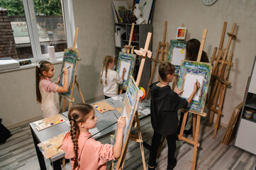 A group of school children with brushes and an easel in art lesson began drawing landscapes. A...