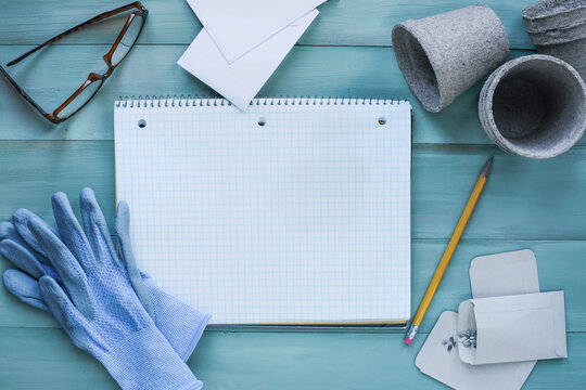 Blank gardening grid notebook surrounded by garden gloves, seeds, pencil, glasses, envelopes and peat pots on a rustic wooden table. Table top view.