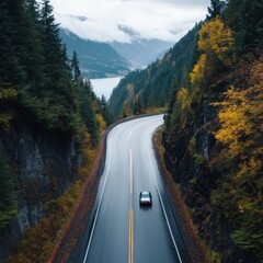 Fototapeta premium A single car drives on a winding mountain road surrounded by lush green and autumnal trees under an overcast sky
