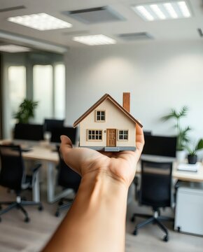 a person is holding a miniature model of a house in an office setting, symbolizing property and investment