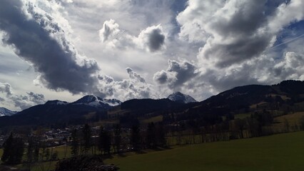Mountain landscape with dark silhouettes, snow-covered peaks, and a dramatic sky filled with textured clouds. A small village is visible below, surrounded by green fields and trees, under dynamic ligh