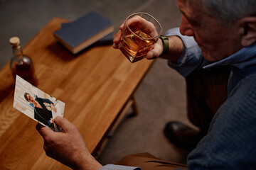 Senior man holding glass of drink and looking at a photograph at wooden table, creating nostalgic...