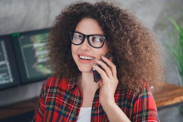 Confident young female programmer talking on smartphone while working on coding project in casual office environment with multiple monitors