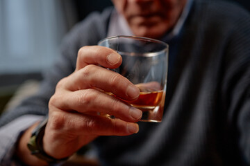 Close-up of elderly person's hand holding glass of whiskey. Warm ambiance created with cozy interior in background