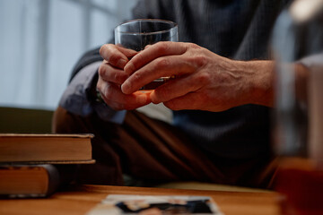 Elderly hands gently cradling glass of water while sitting at table, stack of books and small photograph in background