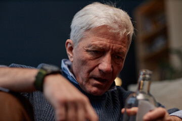 Elderly man deeply reflecting, sitting indoors with bottle in hand wearing blue sweater expressing...