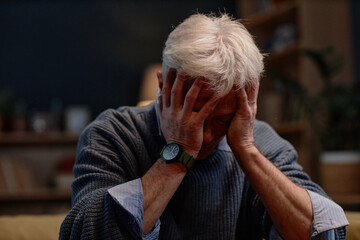 Elderly man covering face with hands while sitting indoors, expressing sadness and stress, wearing...