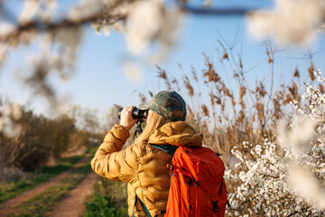 Birdwatching in natural habitat. Woman ornithologist with binoculars observes birds arriving in spring in blooming nature
