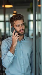 A stylish young man with a well-groomed beard is engaged in a phone conversation, exuding confidence and professionalism. The contemporary office setting, with its sleek design and soft lighting