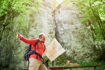 Bearded tourist man studies map while stands in front of large rock in dense forest. Traveler with grey backpack and glasses, pointing ahead, planning outdoor adventure, hiking or climbing route.
