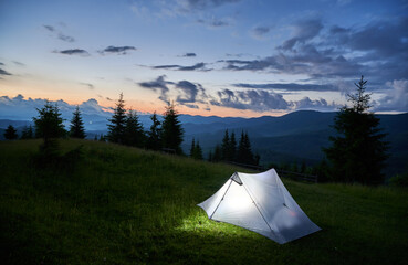 Touristic tent glows warmly on grassy hillside at dusk, surrounded by dark silhouettes of mountains...
