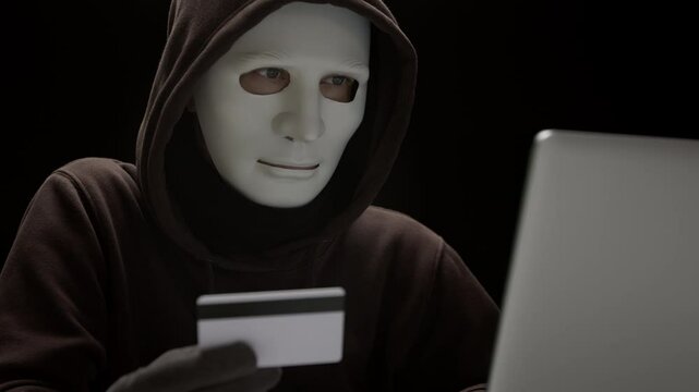 A masked hacker holds a credit card and looks at a laptop. Cybercrime concept.