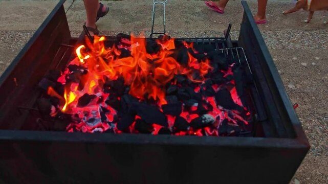 A charcoal barbecue with flames of intense fire and feet of unrecognizable people in flip-flops approaching with a small dog. BEV close up slow motion